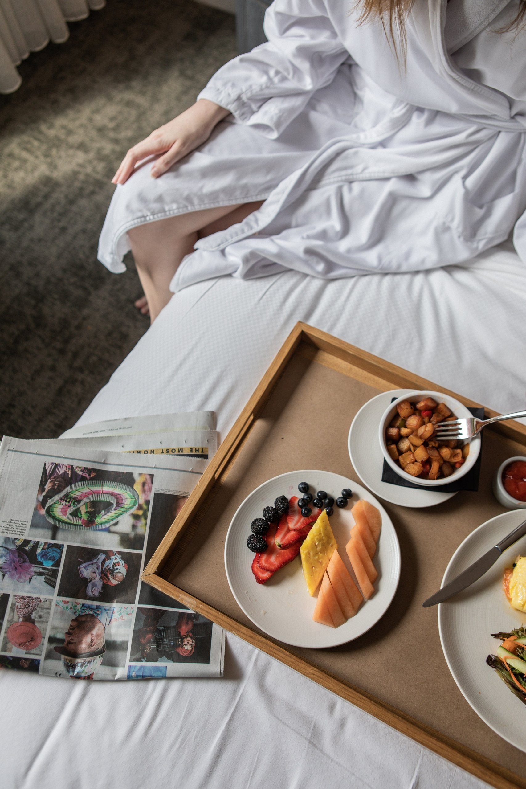 A woman in a robe sitting on a bed with a tray of food, enjoying room service in a Tribeca Hotel