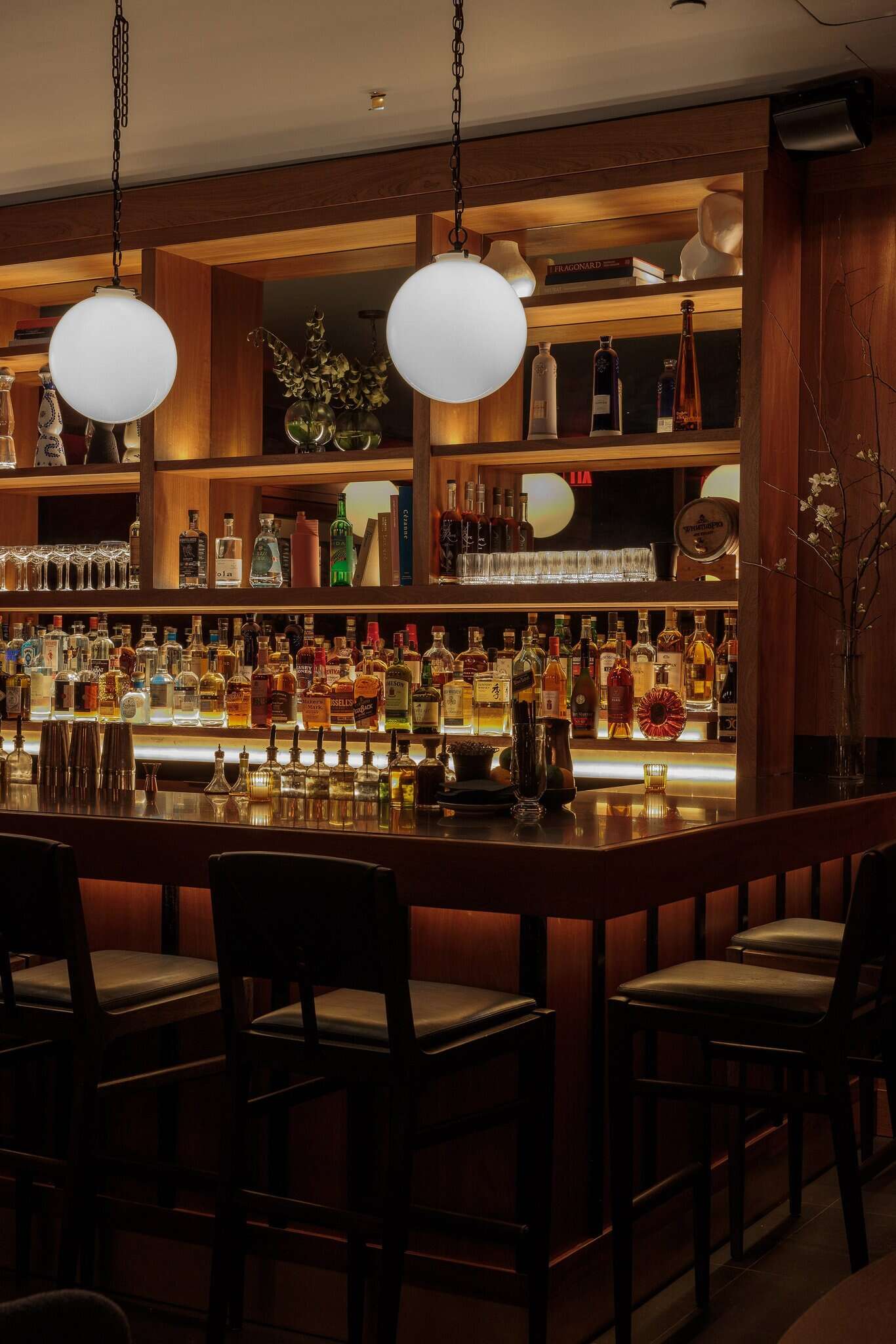 A dimly lit bar with a long counter, stools, and shelves stocked with liquor and glasses.
