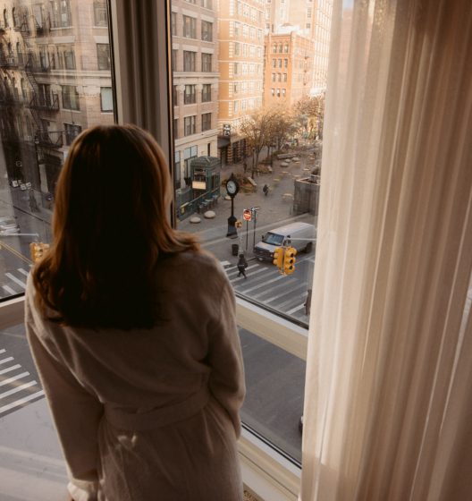 Person in a robe standing at a window, looking out over a city street with crosswalk and traffic lights outside.