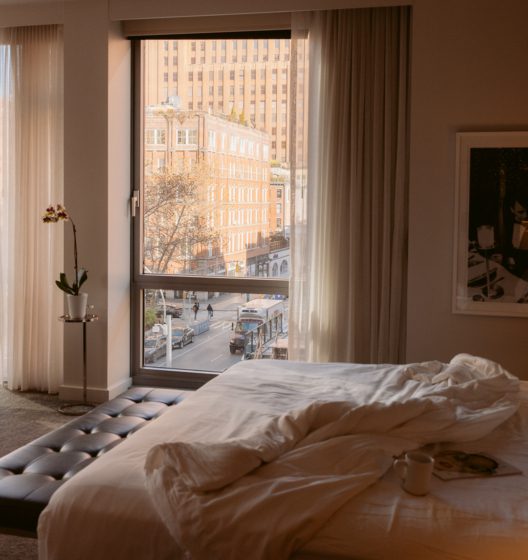 A cozy hotel bedroom with an unmade white duvet on a tufted bed and a large window overlooking a city street bathed in warm light.