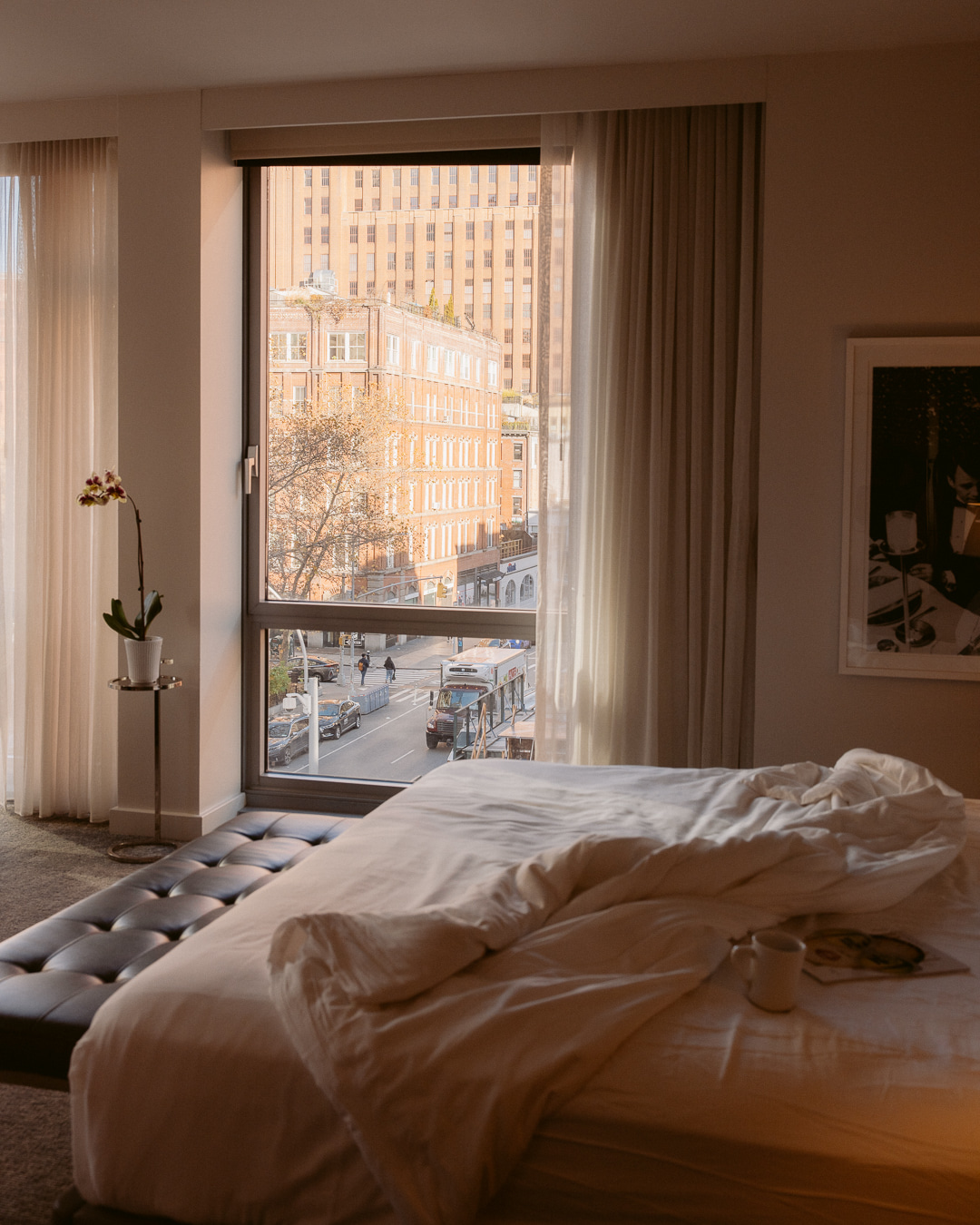 A cozy hotel bedroom with an unmade white duvet on a tufted bed and a large window overlooking a city street bathed in warm light.