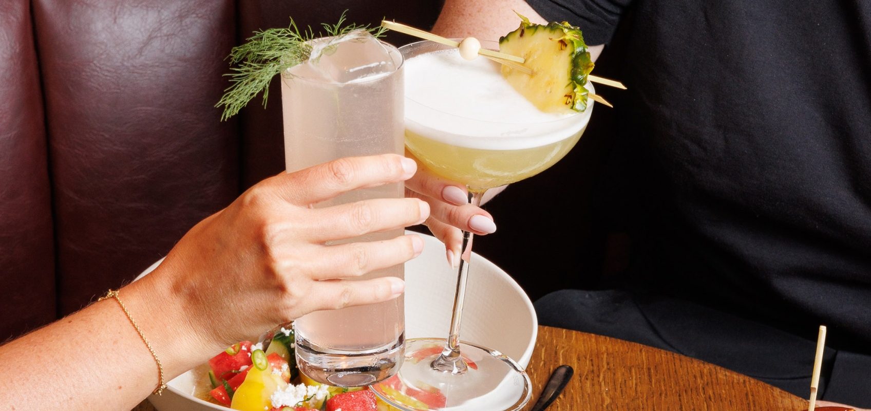 Two people clinking cocktails at a table with food